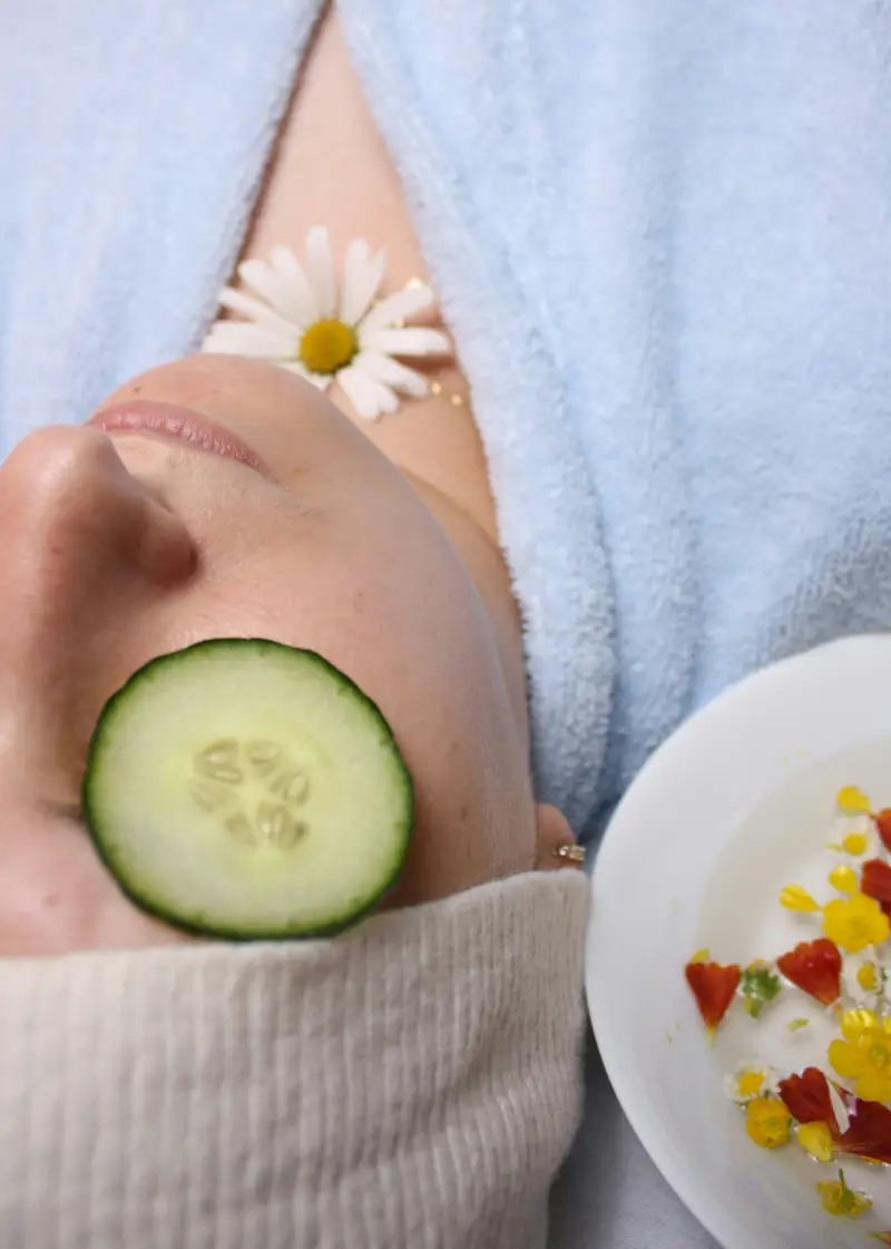 A person relaxing with a cucumber slice on their eye, a daisy on their chest, and a plate of colorful flower petals beside them