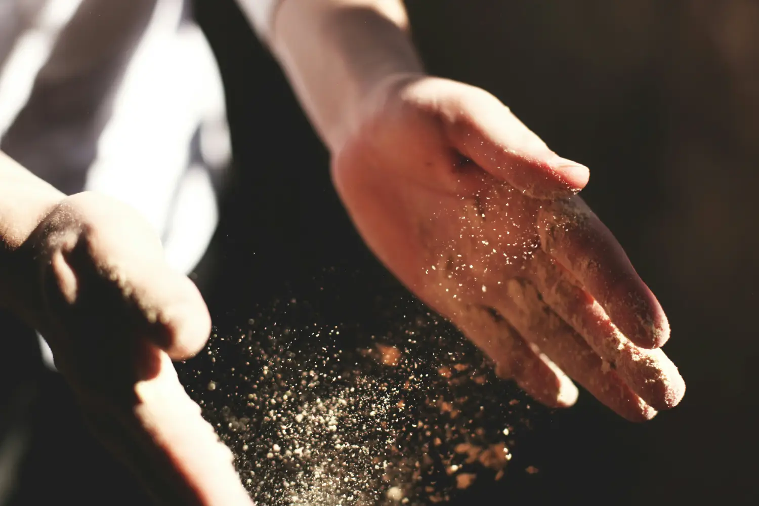 A person’s hands create a cloud of dust as they rub together, with sunlight illuminating the particles floating in the air