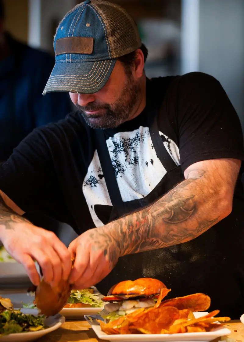 A tattooed chef prepares a plate with a burger, chips, and salad in a lively kitchen setting, showcasing culinary skills and creativity